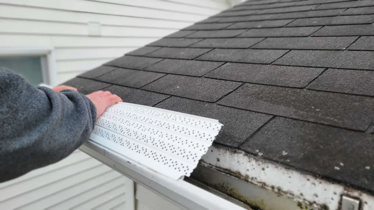Hand sliding a perforated guard onto a gutter below a shingle roof