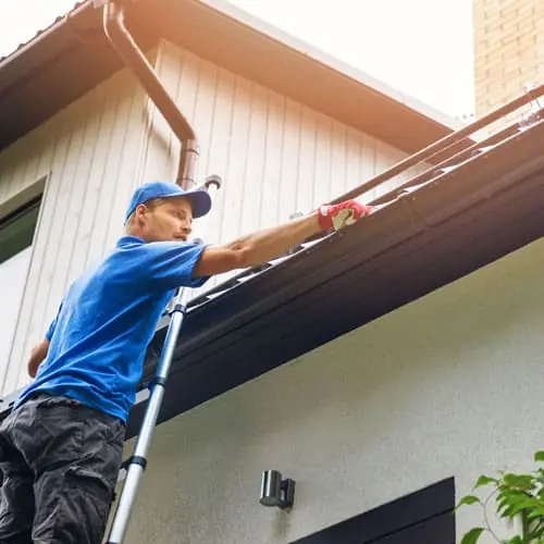 Gutter technician on a ladder checking a dark gutter on a two-story house